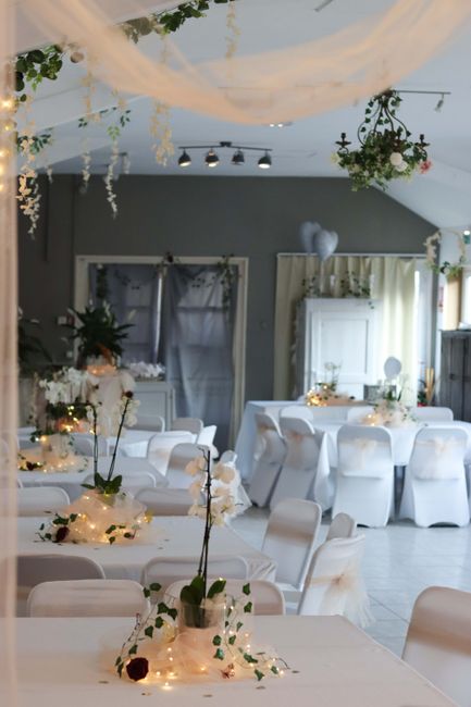 A floral arrangement featuring tall white orchids with dark red centers, placed in a transparent glass vase on a table decorated with warm string lights and white fabric. The background shows a large