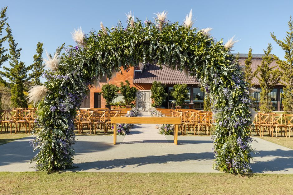 A large floral arch decorated with an abundance of fresh flowers, including white pampas grass, purple lavender, and various greenery, arranged in a lush, cascading style. The arch is situated outdoor