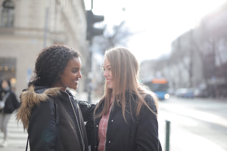 Two women standing outdoors on a city street, smiling and facing each other warmly. They are dressed in casual jackets; one has a dark coat with a fur-lined hood, and the other wears a black jacket ov