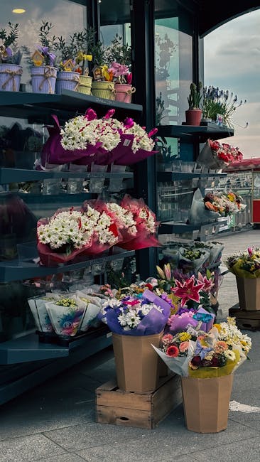 A display of fresh floral arrangements and bouquets on metal shelving and in baskets outside a flower shop, featuring a variety of vibrant flowers including white daisies, pink lilies, purple irises,