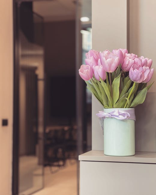 A bouquet of pink tulips with lush green stems and leaves arranged in a pale green, cylindrical ceramic vase tied with a light purple ribbon. The vase is placed on a white surface near a wall, with na