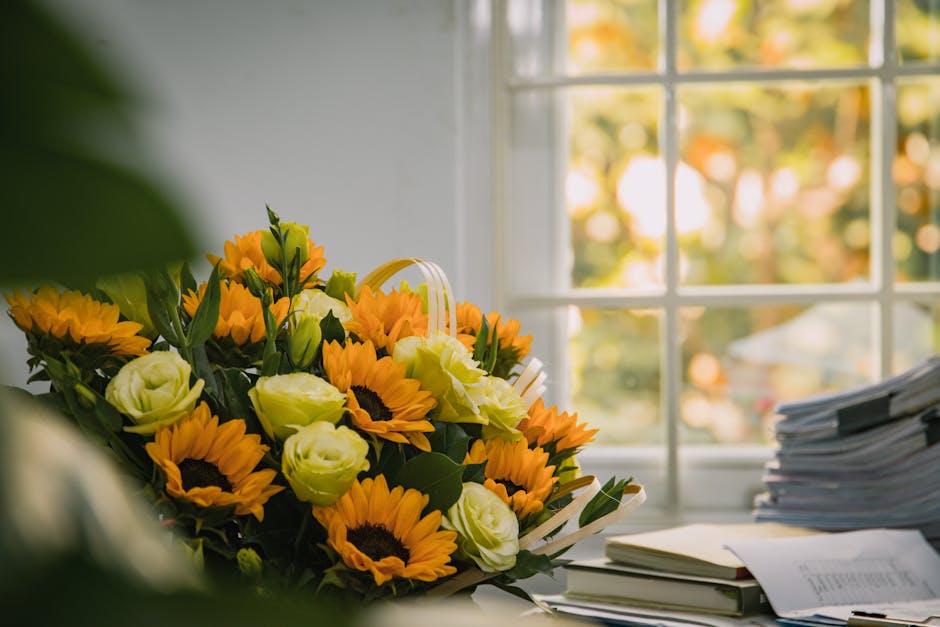 A vibrant floral arrangement featuring bright yellow sunflowers, creamy white Lisianthus, and green foliage placed on a cluttered office desk next to neatly stacked papers and documents. The bouquet i