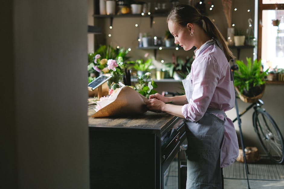A young female florist in a pastel pink shirt and grey apron arranges a bouquet of fresh flowers on a rustic wooden counter inside a well-lit flower shop. The bouquet includes pink roses, white lilies