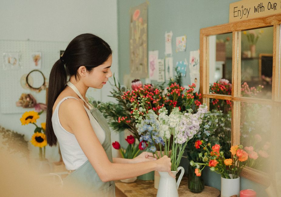 A young woman with dark hair tied back in a ponytail, wearing a grey apron over a white sleeveless top, arranges a bouquet of mixed flowers in a white ceramic pitcher inside a flower shop. The bouquet