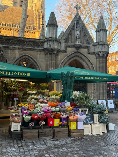 An outdoor flower stall situated in front of a historic gothic-style stone building with pointed arches, turrets, and detailed carvings, featuring a green canopy bearing the website name 'boughtonflow