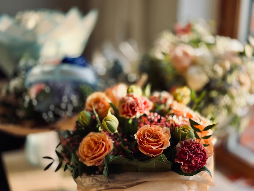 A close-up view of a vibrant floral arrangement featuring primarily peach and deep pink roses, accented with small green foliage and pink buds, arranged in a bouquet with wrapped paper exterior. The b